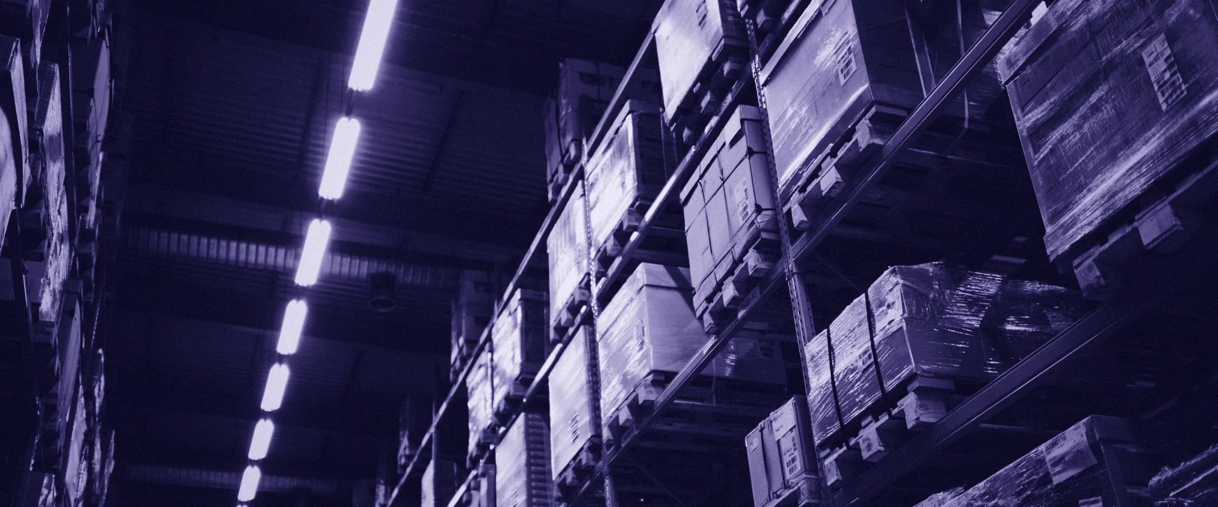 Tall warehouse racks loaded with pallets of cardboard boxes, seen from the floor beside a line of ceiling lights.