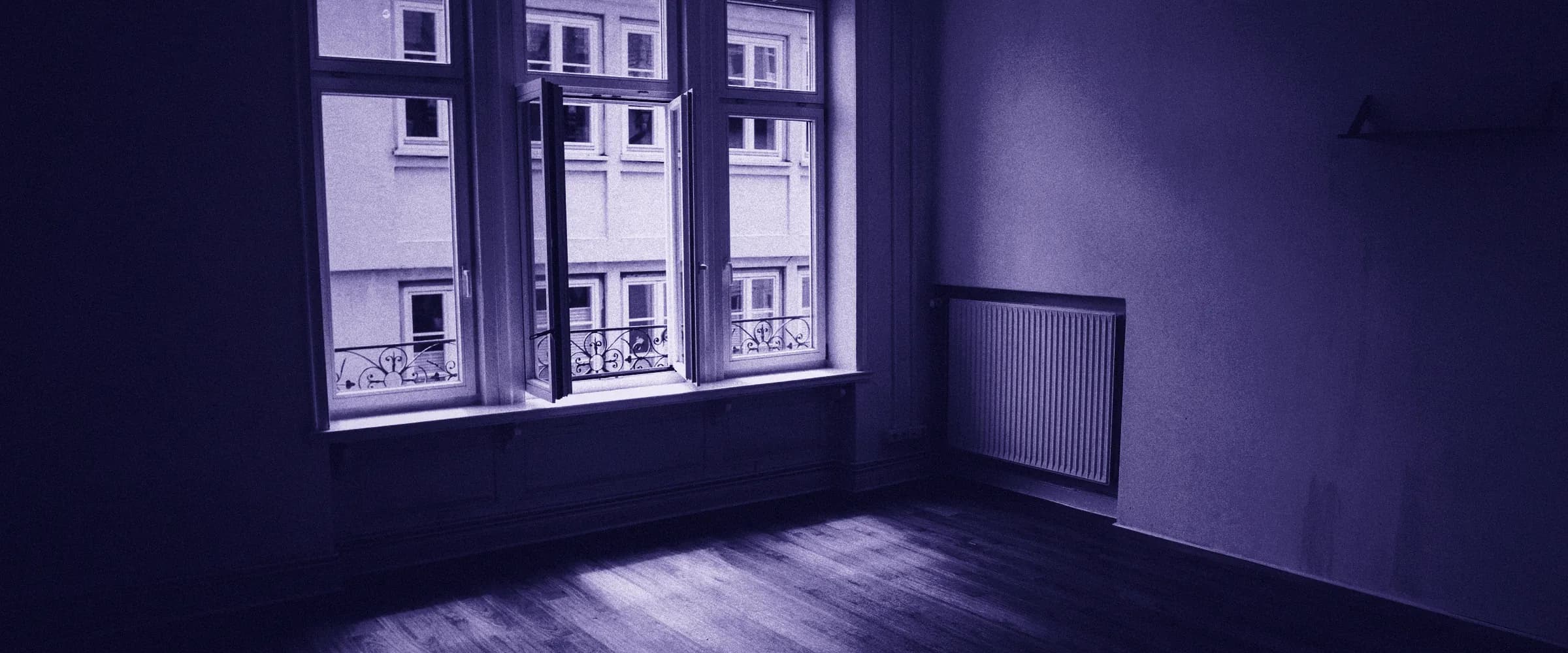 An empty room with a wooden floor, a radiator on the bare wall and a tall window with one casement open onto the building across the street.