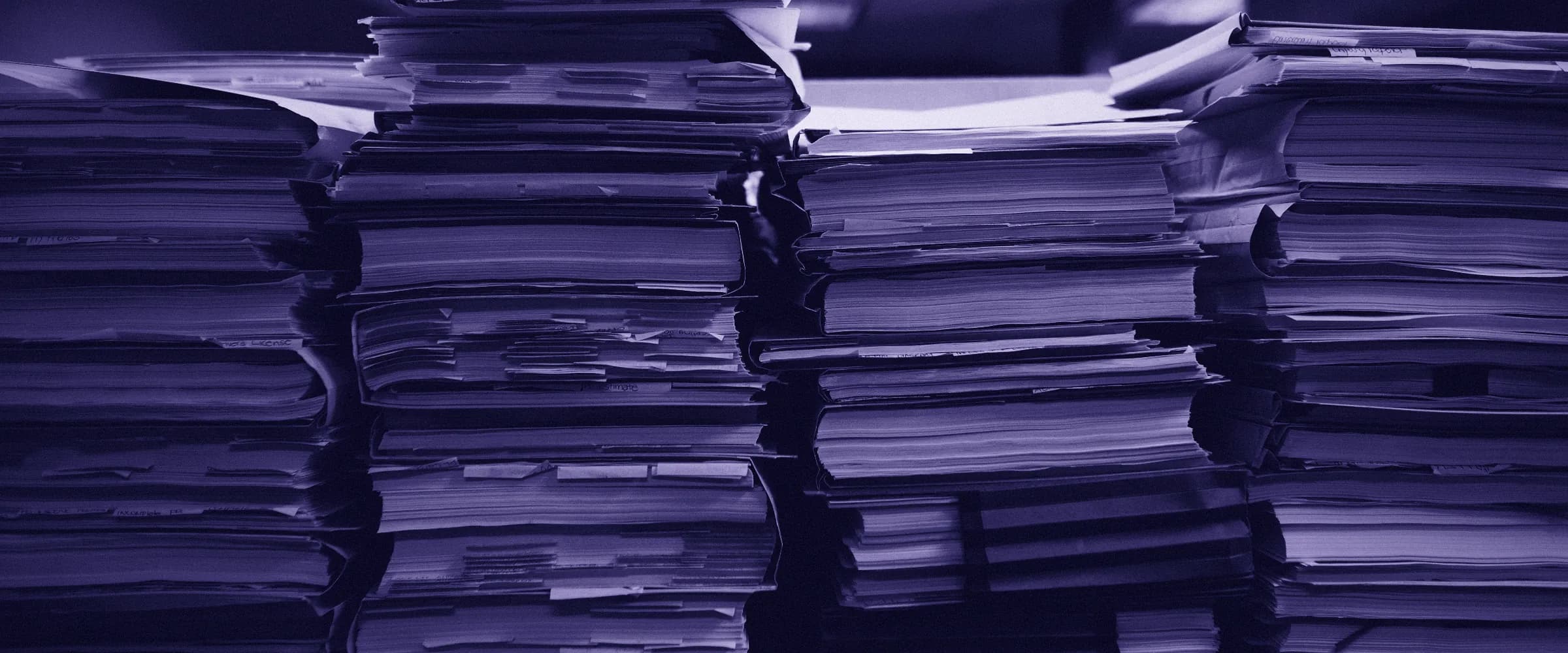 Tall stacks of paper files and folders, with loose pages and tabs sticking out at their edges.