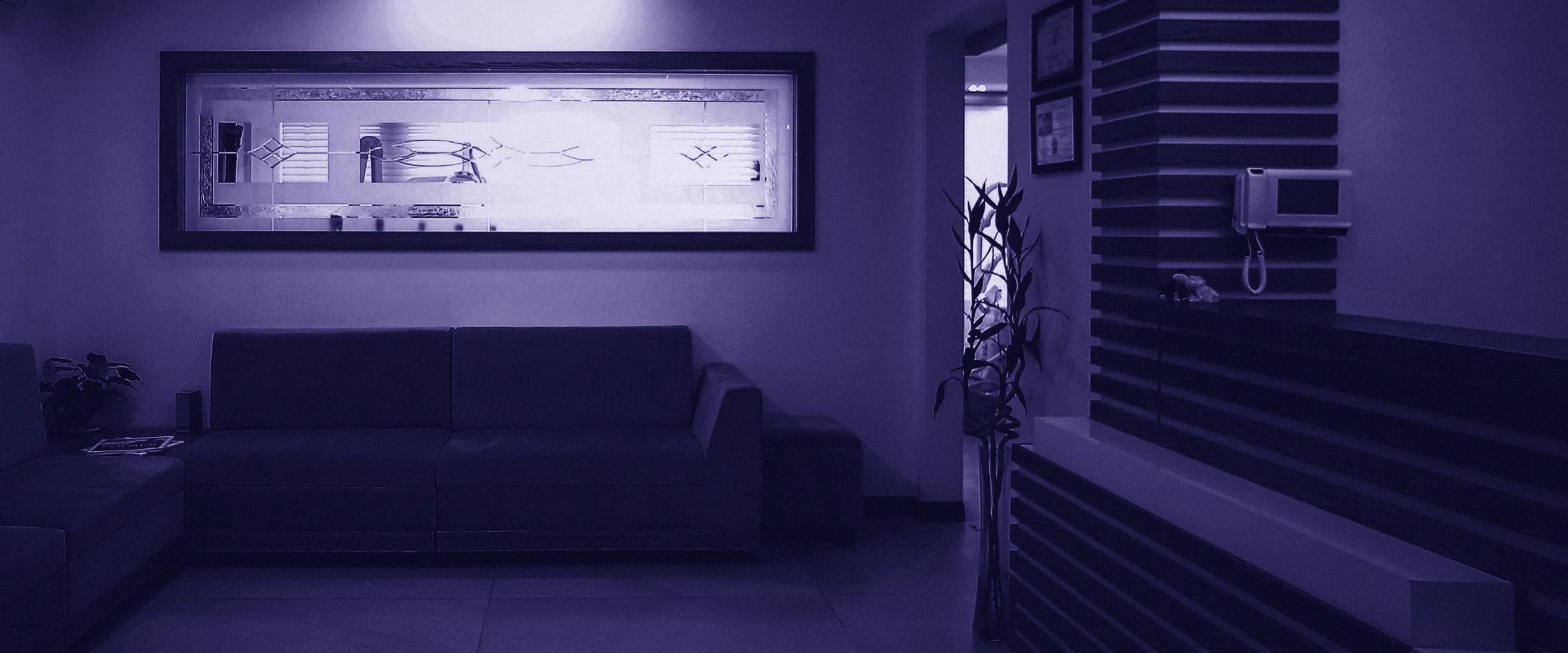A clinic waiting room with a long sofa under a framed frosted-glass panel, and a slatted front desk with a wall phone on the right.