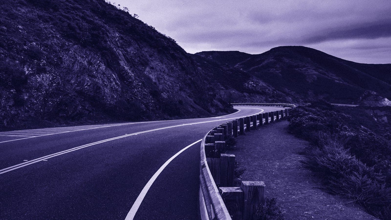A two-lane road curving around a hillside, with a metal guardrail on wooden posts along its outer edge.