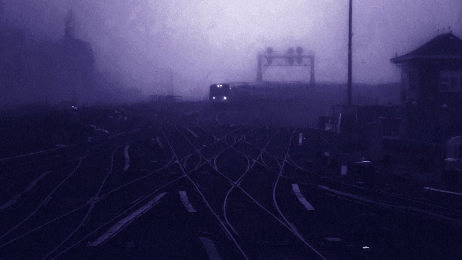 Railway tracks splitting and crossing in a wide rail yard in thick fog, with a signal gantry and a train's headlights far off.