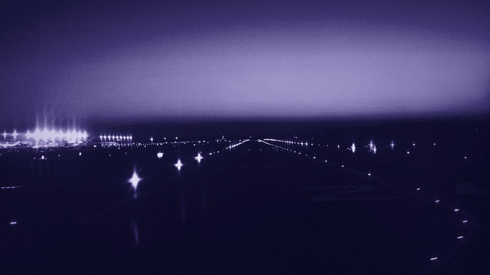An airport runway at dusk, its rows of edge and approach lights running toward a pale band of sky.