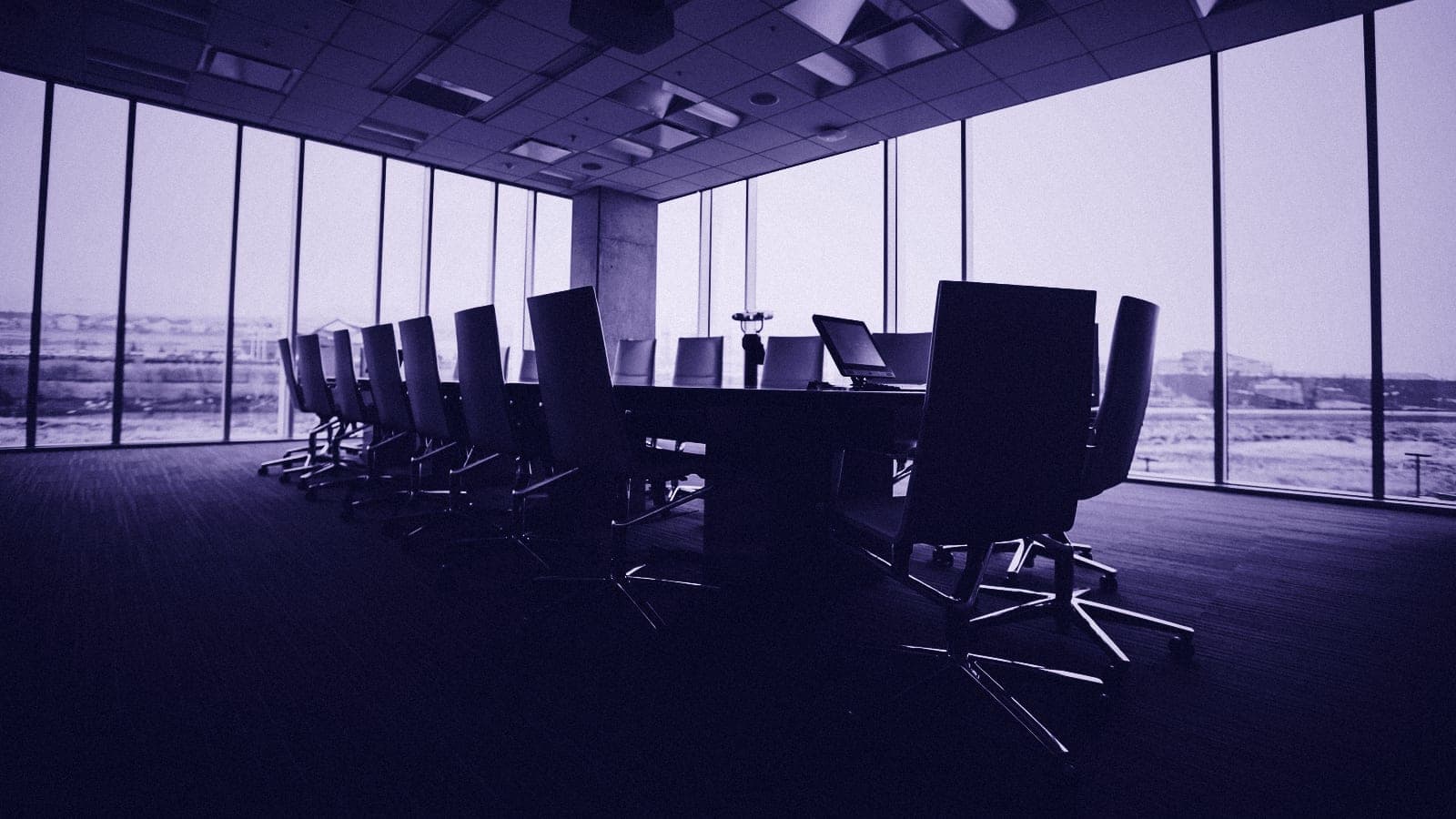 An empty conference room with a long table and high-backed chairs in front of floor-to-ceiling windows.